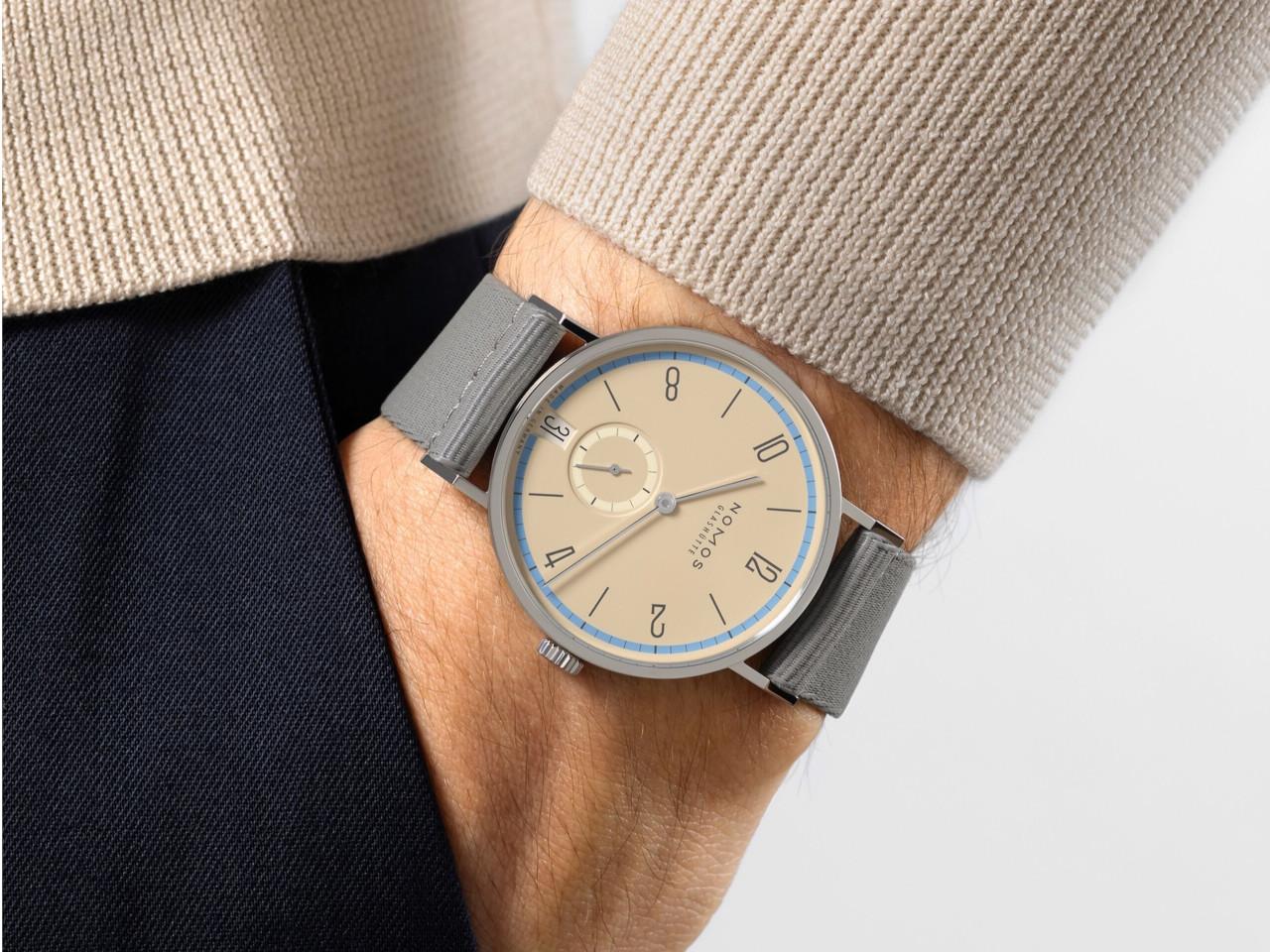 Nomos Glashütte Tangente 38 Datum "175 Years Watchmaking - Ariel" Handaufzug Edelstahl sandfarbenes Zifferblatt graues Textilband Glasboden – Herrenuhren Uhr mit Datum aus Edelstahl. Das Gehäuse ist rund und silber. Das Textilarmband ist grau. Das Zifferblatt ist arabisch Index und beige. – Ansicht 7